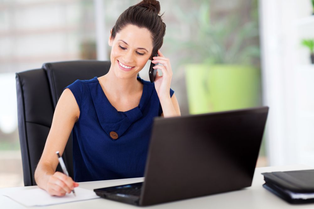 Lady Sales Representative Smiling Talking On The Phone. Writing with a pen on a paper. She has her laptop open on her white desk.